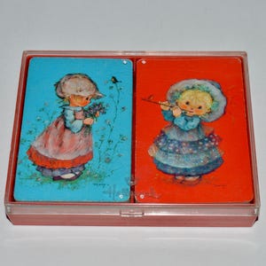 May include: A vintage playing card set featuring two cards with illustrations of children. One card has a girl holding flowers on a blue background, and the other has a girl playing a flute on a red background. The cards are in a pink plastic case.