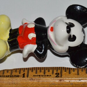 May include: A vintage Mickey Mouse figurine, lying on its side. The figure is primarily black, white, red, and yellow. The figurine is approximately 3 inches long, with a wooden ruler in the foreground.
