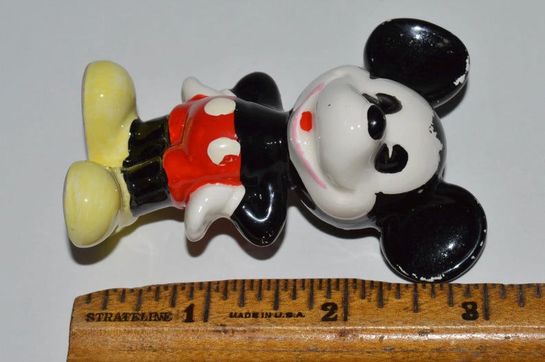 May include: A vintage Mickey Mouse figurine, lying on its side. The figure is primarily black, white, red, and yellow. The figurine is approximately 3 inches long, with a wooden ruler in the foreground.