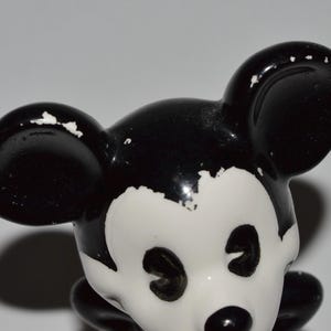 May include: A vintage Mickey Mouse figurine, featuring a black and white color scheme. The figurine's ears, head, and body are black, while the face is white. The paint shows signs of wear, adding to its antique charm.
