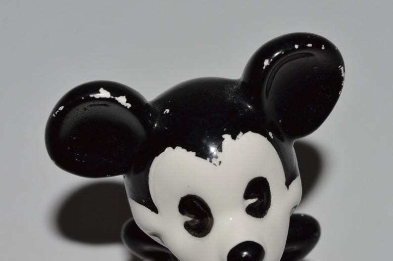May include: A vintage Mickey Mouse figurine, featuring a black and white color scheme. The figurine's ears, head, and body are black, while the face is white. The paint shows signs of wear, adding to its antique charm.