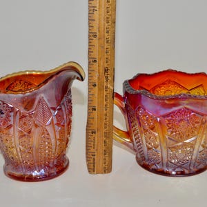 May include: A set of two vintage iridescent glass creamer and sugar bowl. The creamer has a handle and the sugar bowl has two handles. Both pieces are decorated with a geometric pattern and have a red, orange, and yellow color scheme.