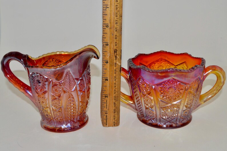 May include: A set of two vintage iridescent glass creamer and sugar bowl. The creamer has a handle and the sugar bowl has two handles. Both pieces are decorated with a geometric pattern and have a red, orange, and yellow color scheme.