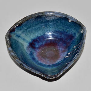 May include: Handmade ceramic bowl with a unique, irregular shape. The interior features a blend of blue, teal, and purple hues, creating a watercolor effect. The rim has a dark, textured edge. A decorative piece for serving or display.
