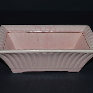 May include: A pale pink ceramic planter with a rectangular shape and a fluted design. The planter has a slightly flared rim and a smooth, glossy finish.