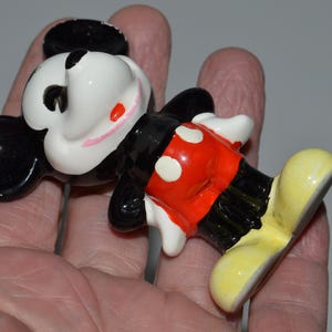 May include: A ceramic figurine of Mickey Mouse, featuring a black head and ears, a white face with red cheeks, a black body, red shorts with white buttons, and yellow shoes. The figurine is a classic Disney collectible.