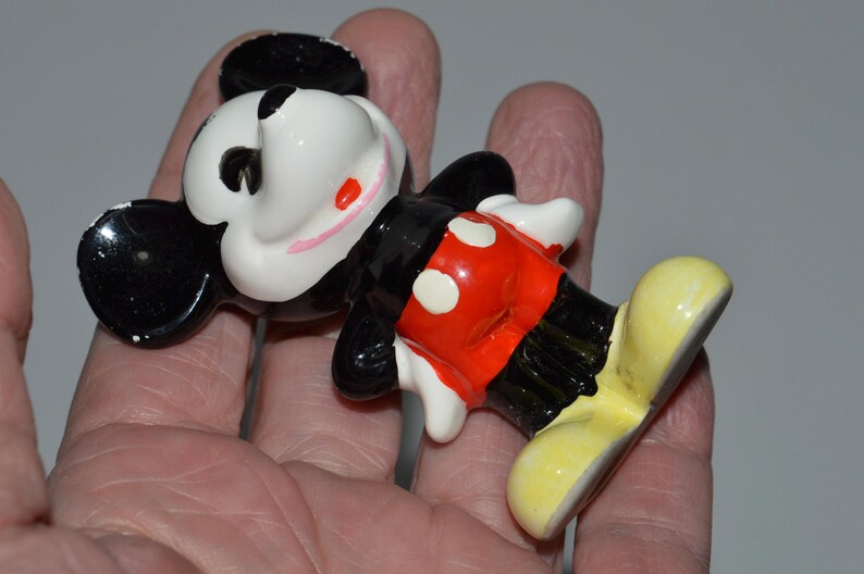May include: A ceramic figurine of Mickey Mouse, featuring a black head and ears, a white face with red cheeks, a black body, red shorts with white buttons, and yellow shoes. The figurine is a classic Disney collectible.