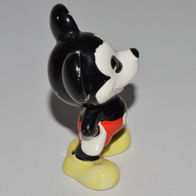 May include: A vintage Mickey Mouse figurine. The figure is primarily black and white, with red shorts and yellow shoes. The figurine is standing and has a classic cartoon appearance.