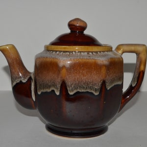 May include: A brown and cream glazed ceramic teapot with a rounded body and a lid. The teapot has a brown handle and a spout.