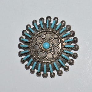 May include: A silver and turquoise brooch with a circular design. The center of the brooch features a turquoise stone surrounded by smaller turquoise stones. The outer edge of the brooch is decorated with silver beads and turquoise stones.