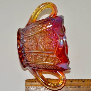 May include: A red and amber glass sugar bowl with a geometric pattern and two handles. The bowl is made of iridescent glass and has a vintage look.
