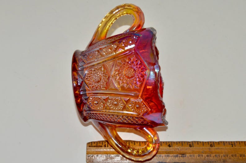 May include: A red and amber glass sugar bowl with a geometric pattern and two handles. The bowl is made of iridescent glass and has a vintage look.