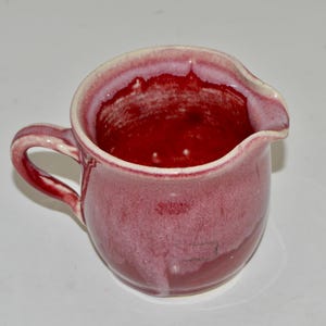 May include: A small, red ceramic creamer with a white rim and a white glaze on the outside. The creamer has a handle and a spout.