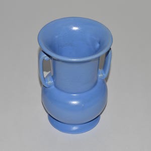 May include: A light blue ceramic vase with two handles. The vase has a bulbous body and a flared rim. The vase is approximately 7 inches tall and 5 inches wide.