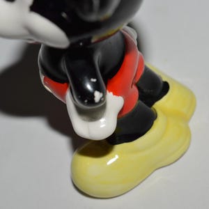May include: A ceramic figurine of Mickey Mouse, featuring black, red, white, and yellow colors. The figure has black legs and arms, red shorts, white gloves, and yellow shoes. The figurine is a collectible item.