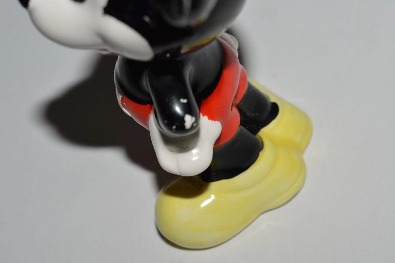 May include: A ceramic figurine of Mickey Mouse, featuring black, red, white, and yellow colors. The figure has black legs and arms, red shorts, white gloves, and yellow shoes. The figurine is a collectible item.