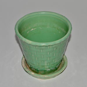 May include: A light green ceramic planter with a basket weave design. The planter sits on a matching saucer. The rim of the planter has a scalloped edge. The planter is empty.
