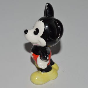May include: A vintage Mickey Mouse figurine. The figure has a black head and body, white face and hands, red shorts, and yellow shoes. The figurine is standing and facing to the side.