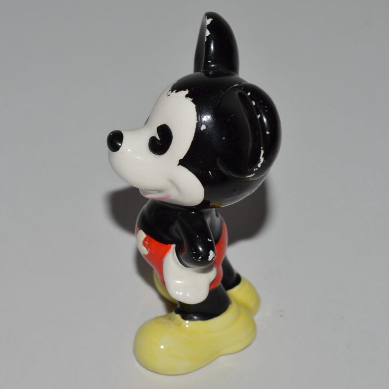 May include: A vintage Mickey Mouse figurine. The figure has a black head and body, white face and hands, red shorts, and yellow shoes. The figurine is standing and facing to the side.