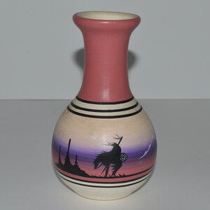 May include: A pink and white ceramic vase with a Native American design. The vase has a round body and a narrow neck. The design features a silhouette of a Native American on horseback, riding into a sunset.