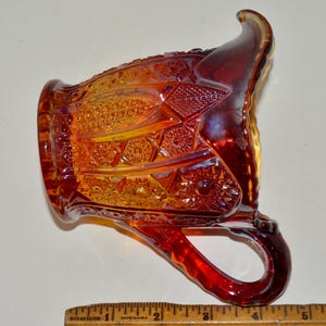 May include: A red and amber glass pitcher with a decorative pattern. The pitcher has a handle and a spout.