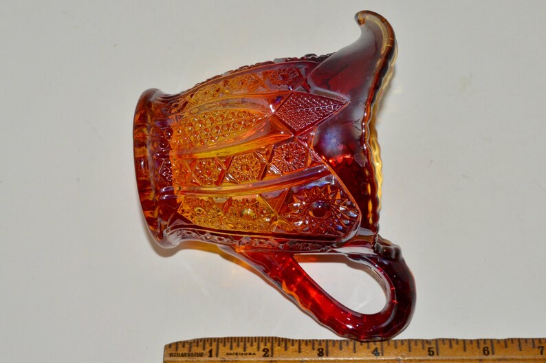 May include: A red and amber glass pitcher with a decorative pattern. The pitcher has a handle and a spout.
