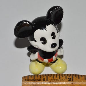 May include: A small, ceramic Mickey Mouse figurine. The figure has a black head and ears, a white face, a black body, red shorts, and yellow shoes. The figurine is standing on a white surface.