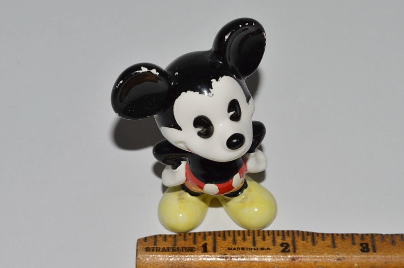 May include: A small, ceramic Mickey Mouse figurine. The figure has a black head and ears, a white face, a black body, red shorts, and yellow shoes. The figurine is standing on a white surface.