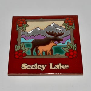 May include: A square ceramic tile with a red border features a scenic illustration. The image depicts a moose and a calf in front of mountains and a lake. The words "Seeley Lake" are at the bottom, with berry and leaf accents.