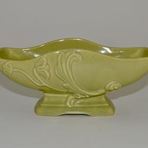 May include: A green ceramic planter with a footed base and an ornate design. The planter is shaped like a bowl with a wide opening and a narrow base.