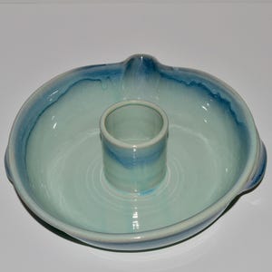 May include: A ceramic chicken roaster with a light blue and turquoise glaze. The round dish has a central cylinder for holding the chicken upright during cooking. The rim and cylinder have a darker blue accent.