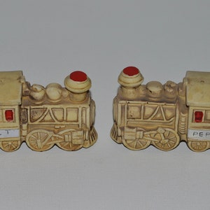 May include: A set of two ceramic salt and pepper shakers shaped like vintage train engines. The shakers are painted in a cream color with brown accents and have red tops. The salt shaker is labeled "SALT" and the pepper shaker is labeled "PEPPER".