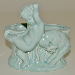 May include: A light blue ceramic planter shaped like a tree trunk with a cherub figure sitting on top. The cherub is holding onto the tree trunk and has its head tilted down.