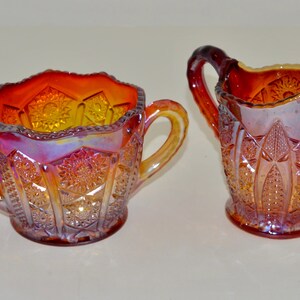 May include: A set of two vintage glass sugar and creamer in a red and orange iridescent carnival glass pattern. The creamer has a handle on the side and the sugar bowl has two handles.