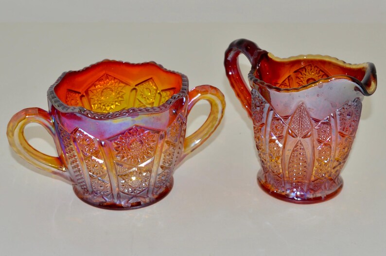 May include: A set of two vintage glass sugar and creamer in a red and orange iridescent carnival glass pattern. The creamer has a handle on the side and the sugar bowl has two handles.