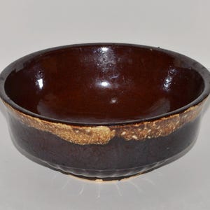 May include: A round, dark brown ceramic bowl with a glossy finish. The rim features a band of lighter, textured tan color. The bowl's exterior has a slightly ridged design near the base, adding visual interest to the piece.