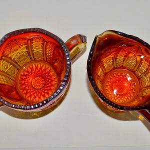 May include: A set of two vintage glass creamer and sugar bowls. The bowls are made of amber glass with a red and orange pattern. The bowls have a scalloped edge and a raised design.
