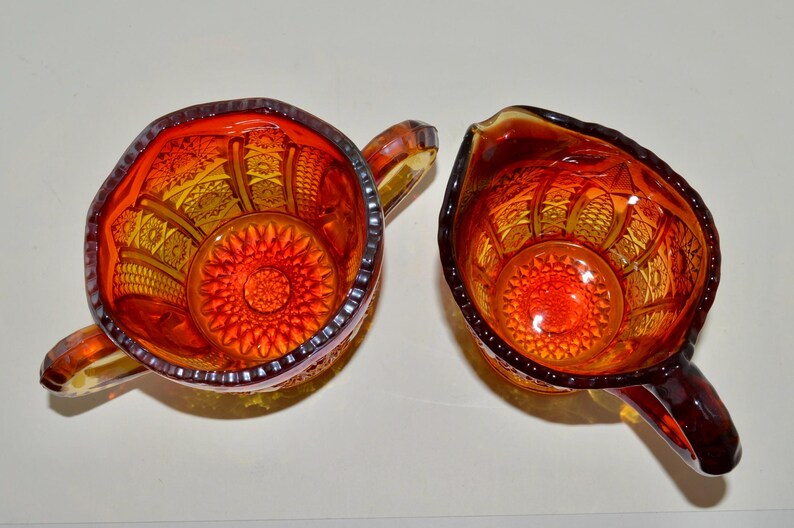 May include: A set of two vintage glass creamer and sugar bowls. The bowls are made of amber glass with a red and orange pattern. The bowls have a scalloped edge and a raised design.