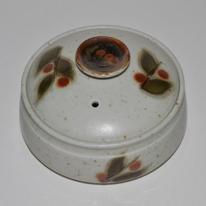 May include: A ceramic container with a lid. The lid features a small, raised, circular handle and is decorated with green and red leaf and berry designs. The container is off-white with brown speckles.