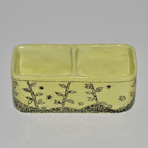 May include: A light yellow ceramic divided dish with a hand-drawn black botanical design. The dish has two compartments and is rectangular with rounded corners. The botanical design includes stylized plants, dots, and swirls.
