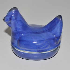 May include: A blue ceramic chicken shaped dish with a lid. The dish is glazed in a light blue color with a darker blue glaze on the chicken's head.