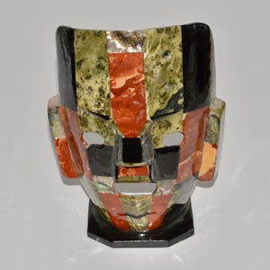 May include: A mosaic mask with a geometric design. The mask features a combination of colors, including black, red, green, and beige. The mask has eye and mouth openings. The base of the mask is black.