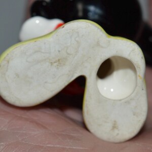 May include: Close-up of the bottom of a vintage ceramic figurine, featuring a yellow and white shoe-like base with a hole. The figurine's black head and white face are visible in the background.