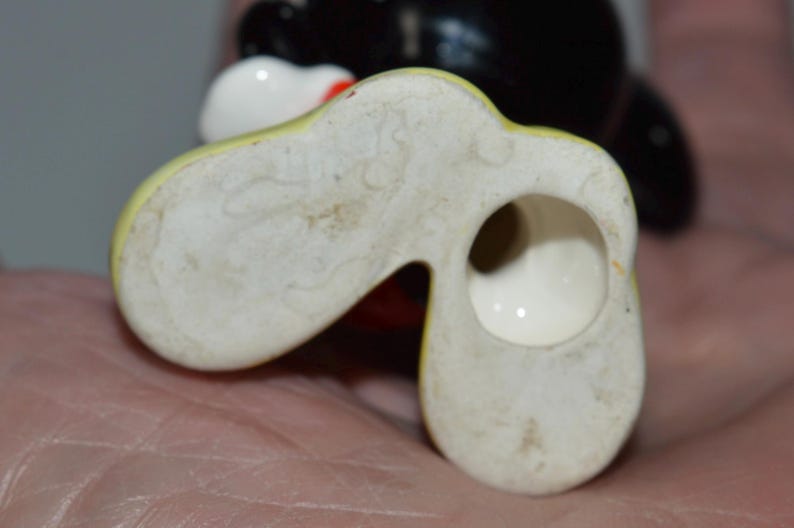 May include: Close-up of the bottom of a vintage ceramic figurine, featuring a yellow and white shoe-like base with a hole. The figurine's black head and white face are visible in the background.