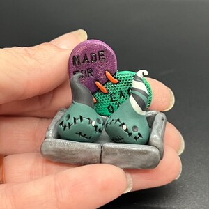 Peut inclure: Deux figurines de fantômes cousues vertes et noires avec des chapeaux sont assises dans une boîte en pierre grise. Une pierre tombale violette avec le texte "Made For Each Other" se trouve derrière les fantômes.