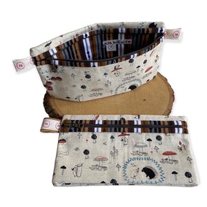 May include: Two zippered pouches with a mushroom and hedgehog print. The pouches are made of beige linen fabric with brown, black, and white stripes on the inside. The larger pouch has a label that reads "Home Made With Love".