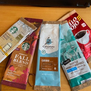 May include: Four different coffee bags with various branding and designs. The bags are all brown and have a cork-like texture. The bags are labeled with the names of the coffee brands, including "Starbucks", "Caribou", and "Seattle Market Inspired".