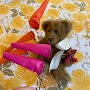 May include: A brown teddy bear wearing a white sash and holding a red, blue, and white toy airplane. The bear is surrounded by four colorful fabric windsocks in shades of orange and pink.