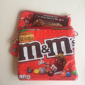 Op de afbeelding: Twee rode etuis met rits met M&M's snoepverpakkingsontwerpen. Eén etui heeft een pindakaas M&M's ontwerp en de andere heeft een verscheidenheid aan M&M's snoepjes. De etuis hebben een hersluitbare rits en een klein sleutelhangeretui.