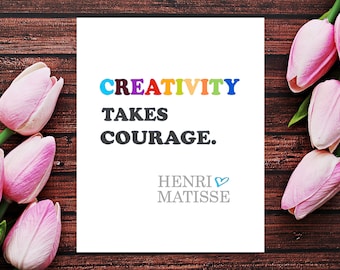 Könnte beinhalten: Eine weiße Karte mit dem Text "CREATIVITY TAKES COURAGE." in bunten Buchstaben. Der Text "HENRI MATISSE" steht darunter in Grau mit einem kleinen blauen Herzsymbol. Die Karte ist von rosa Tulpen auf einem braunen Holzgrund umgeben.